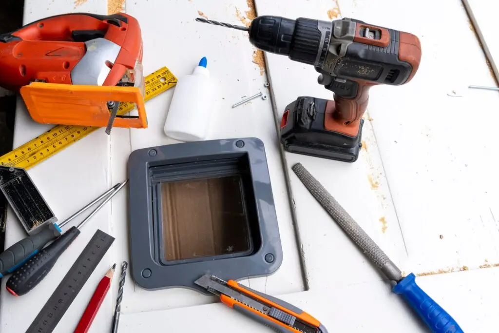Assorted tools on a white wooden work surface: orange cordless drill, tape measure, rulers, and a handheld device with a screen.
