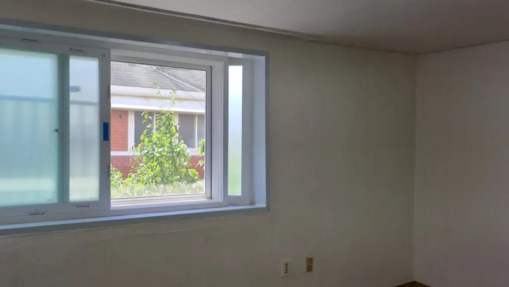 Interior room with a large white-framed window; outside, green foliage and a brick building are visible against a pale wall interior.