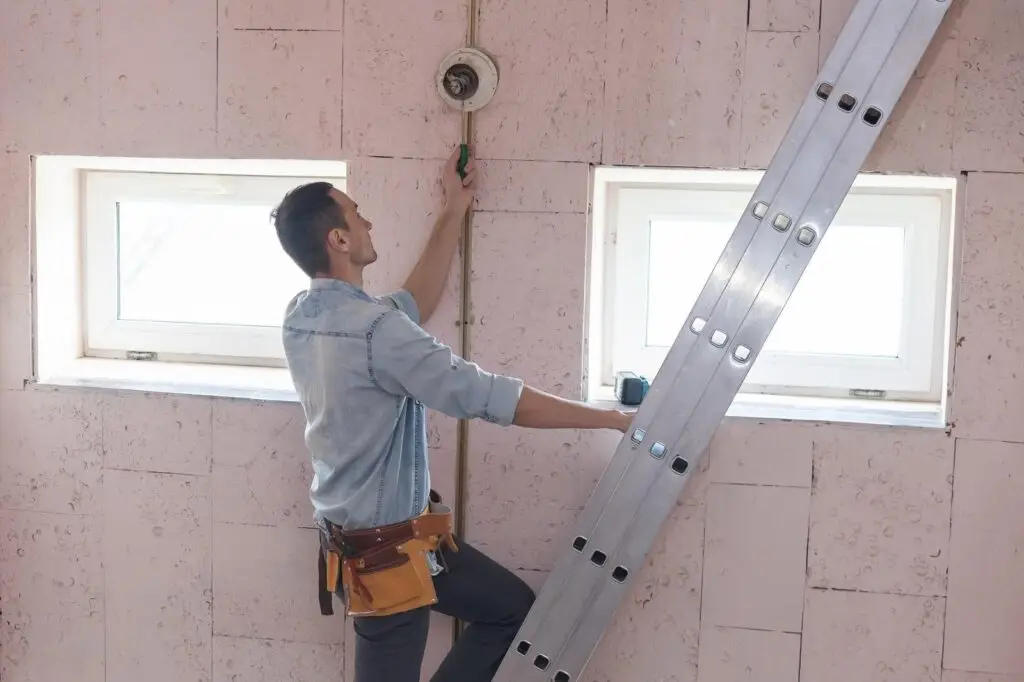 Construction worker standing on a ladder, wiring a wall in a room under renovation, with two windows nearby.